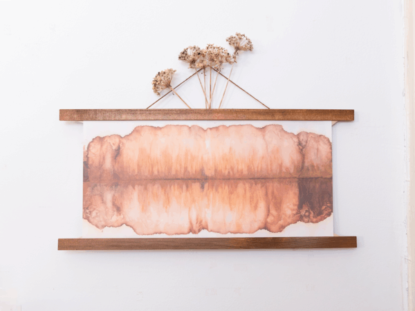 a chromatogram where the top and bottom mirror each other in hazy tans and browns. The print is hung with wooden poster bars at the top and bottom of the print. Dried flowers extend from the top of the frame.